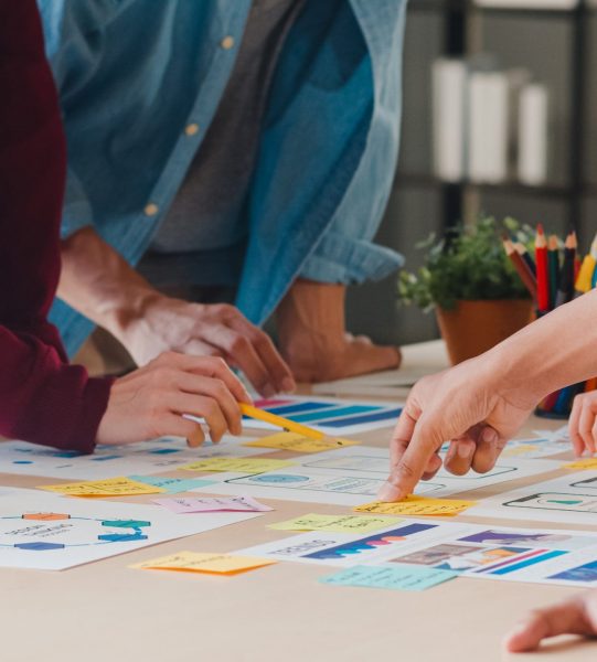 Asian businessmen and businesswomen meeting brainstorming ideas about creative web design planning application and developing template layout for mobile phone project working together in small office.