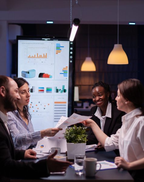 Workaholics businesspeople brainstorming financial company ideas analyzing strategy paperwork late at night in business office meeting room. Multi-ethnic coworkers checking solution
